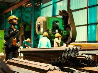 Supervisor in helmet with clipboard standing next to stacked angle-iron profiles
