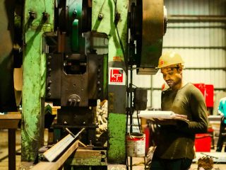 Smiling operator holding a notebook beside a power-press machine