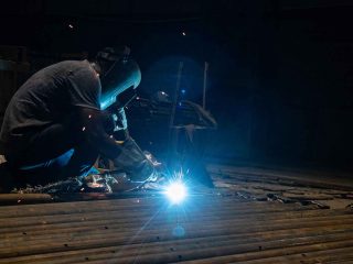 Welder crouched over pipes, bright arc illuminating a dim workshop